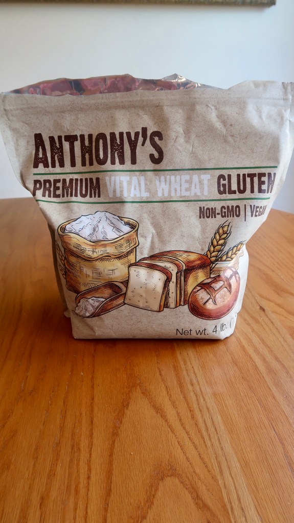 A 4-pound bag of Anthony's Premium Vital Wheat Gluten on a wooden table, featuring illustrations of flour, bread, and wheat, marked as Non-GMO and Vegan.