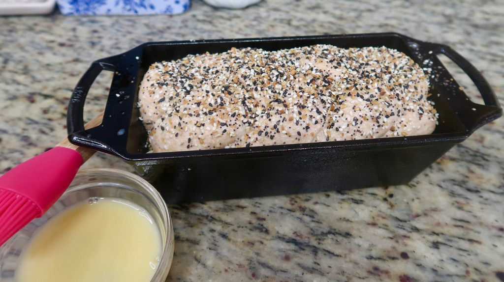 A loaf of dough topped with an assortment of seeds, placed in a black baking dish beside a small bowl filled with liquid, on a granite countertop.
