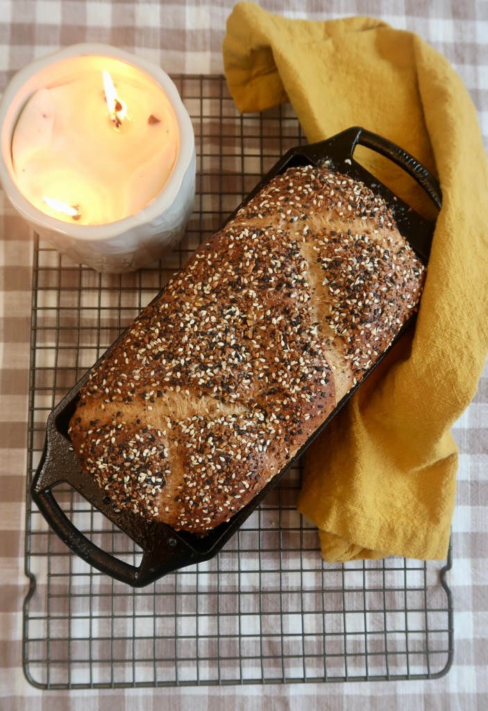 A freshly baked loaf of bread topped with sesame and other seeds, placed on a cooling rack next to a lit candle and a yellow cloth.