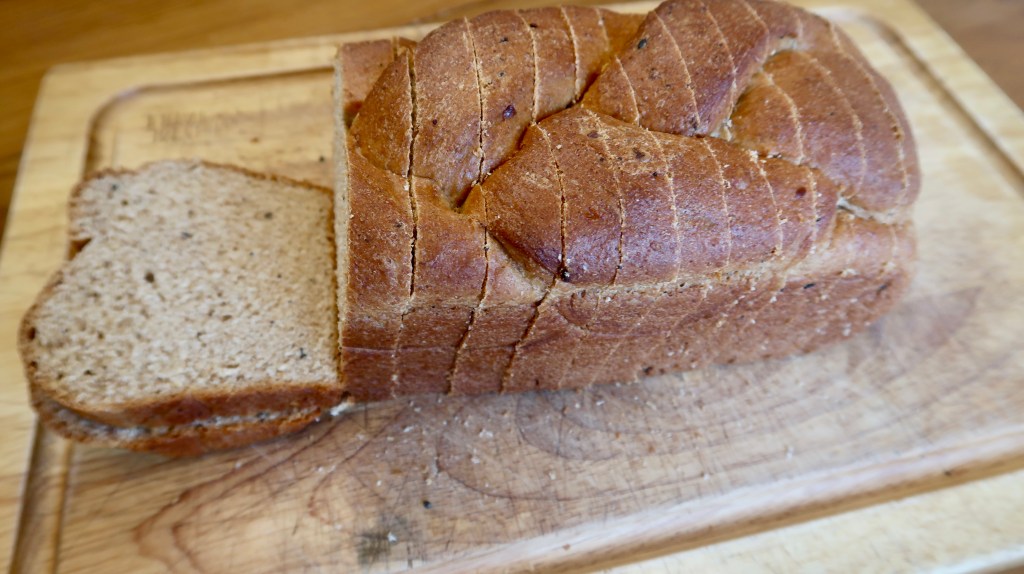 A freshly baked loaf of braided bread on a wooden cutting board, with one slice cut and resting beside the loaf.