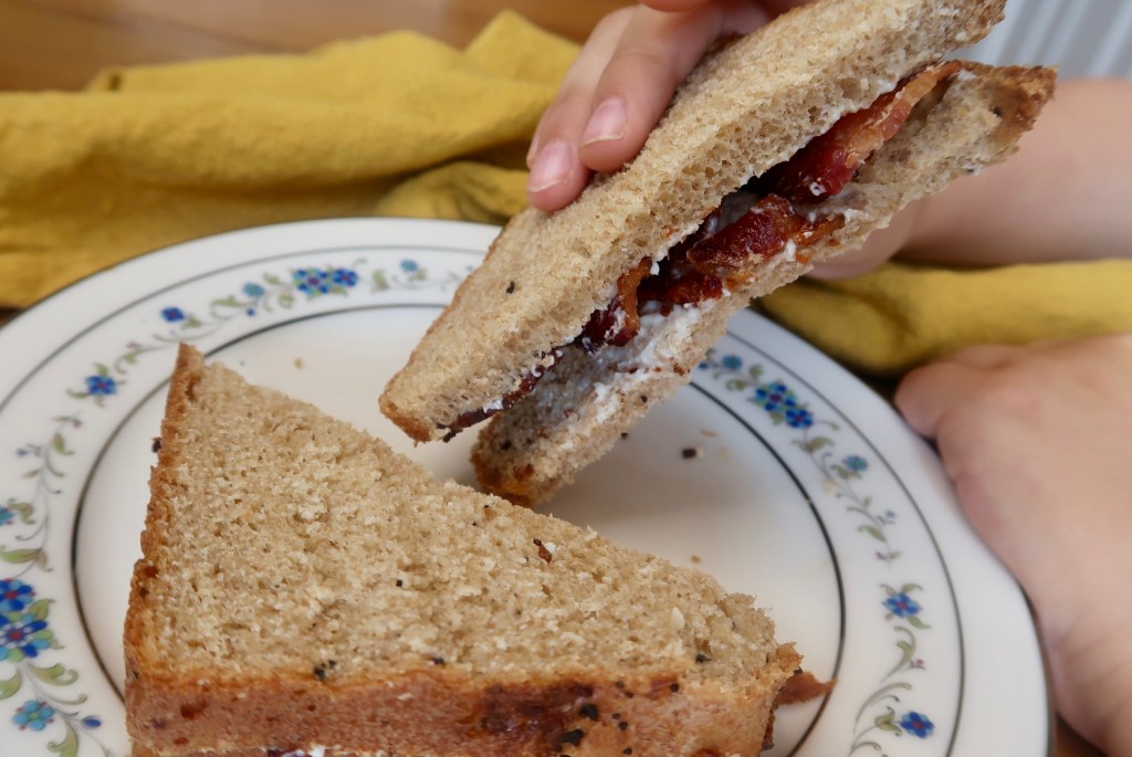A close-up of a person's hand holding a sandwich made with whole grain bread, layered with bacon and cream cheese, placed on a decorative plate with a floral design.