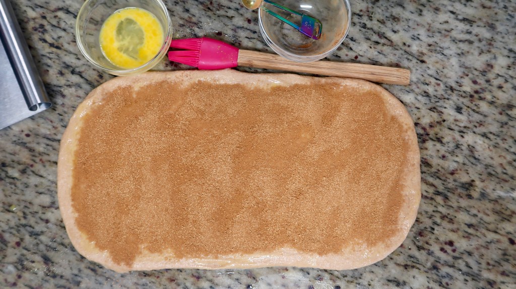 A countertop with rolled dough spread with brown sugar, a bowl of beaten egg, a pink pastry brush, and a wooden rolling pin.