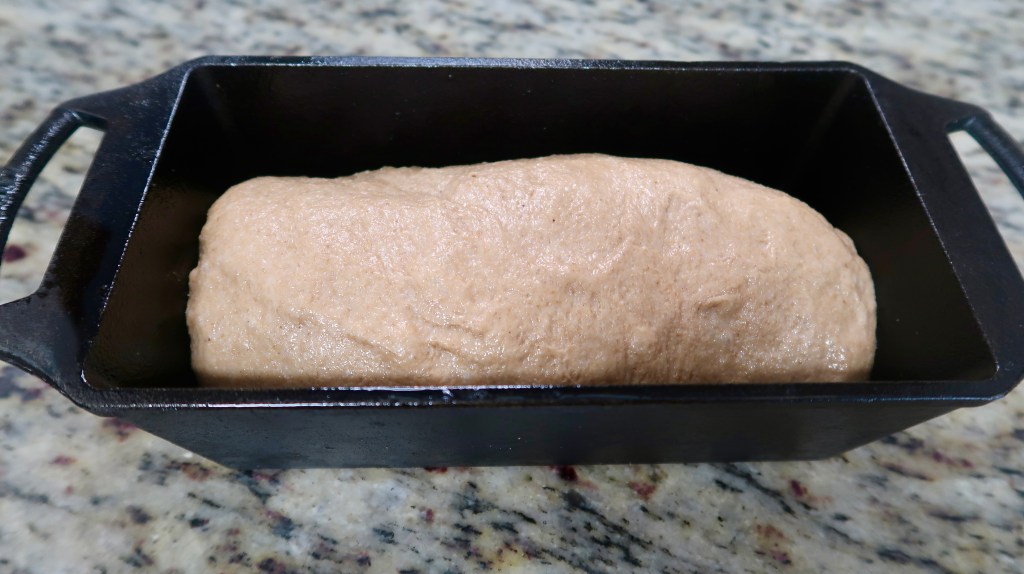 A loaf of dough resting in a black cast iron baking pan on a granite countertop.