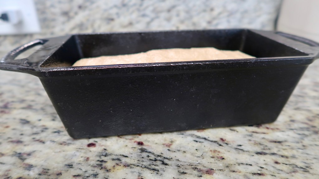 A black cast iron loaf pan with a light brown mixture inside, placed on a granite countertop.