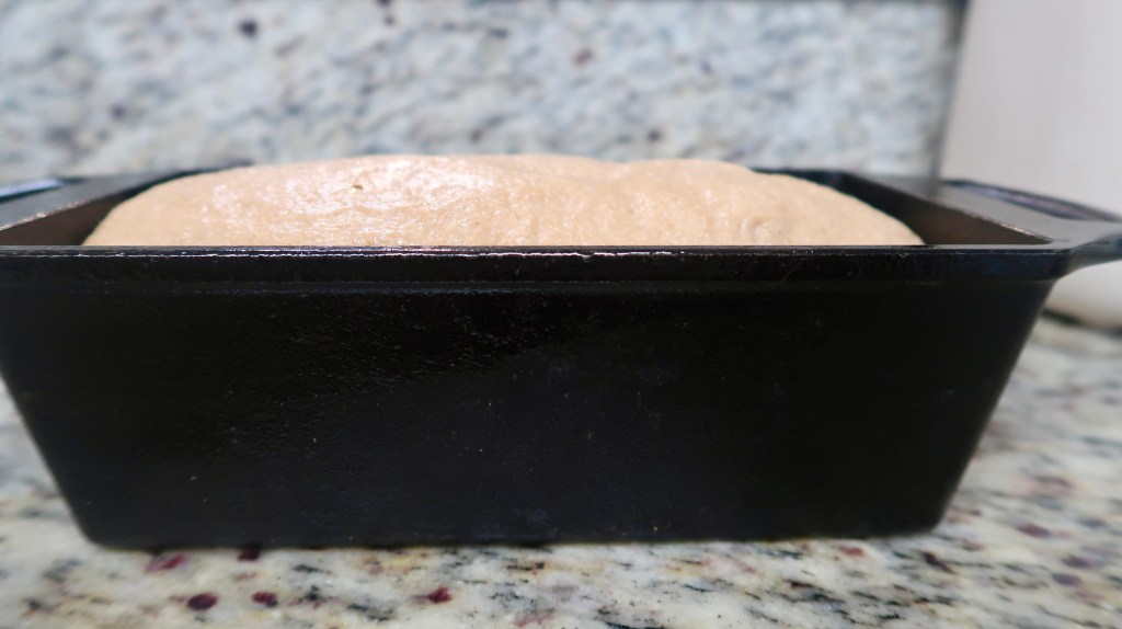 A side view of a loaf of dough rising in a black baking pan on a granite countertop.