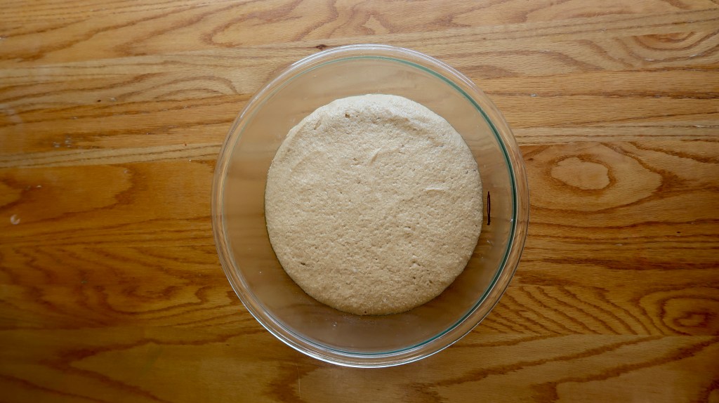 A glass bowl containing a round, risen dough on a wooden surface.