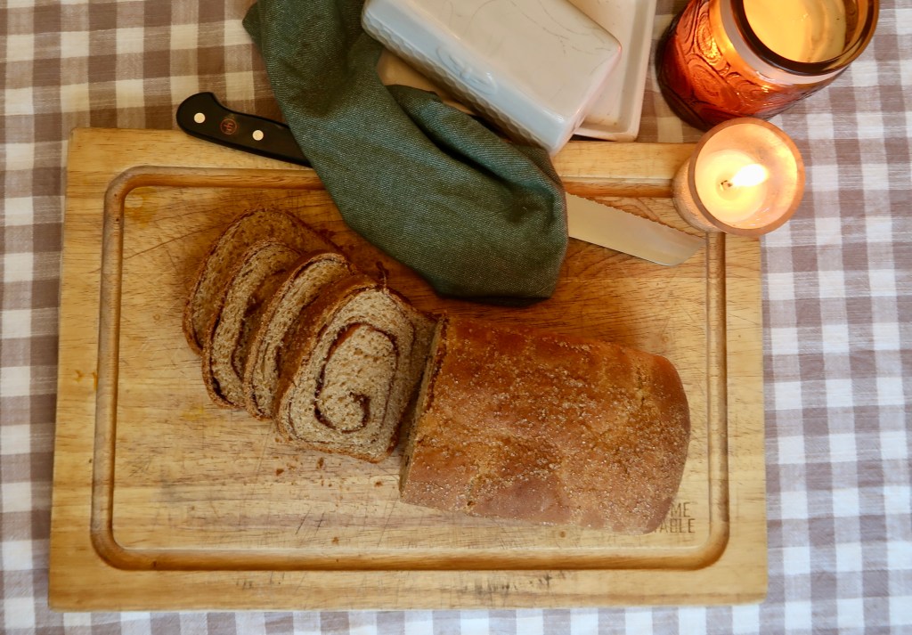A wooden cutting board with a loaf of sliced bread, a knife, a green cloth, a candle, and soap in the background, all set on a checkered tablecloth.