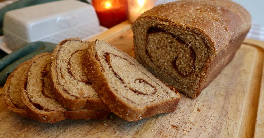 Sliced cinnamon swirl bread on a wooden cutting board, with a whole loaf in the background.