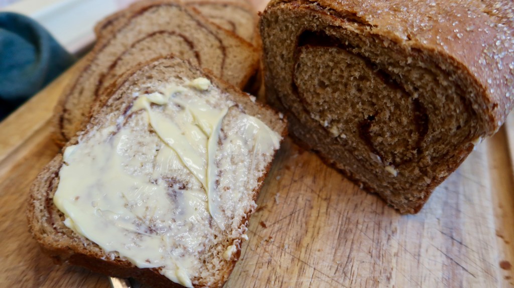 Sliced loaf of cinnamon bread with a slice spread with butter, resting on a wooden cutting board.