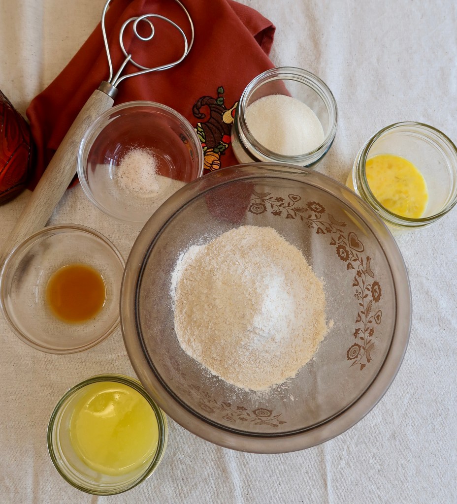 A glass bowl of flour surrounded by small glass containers holding various baking ingredients like sugar, butter, eggs, and vanilla extract, with a whisk and a red cloth in the background.