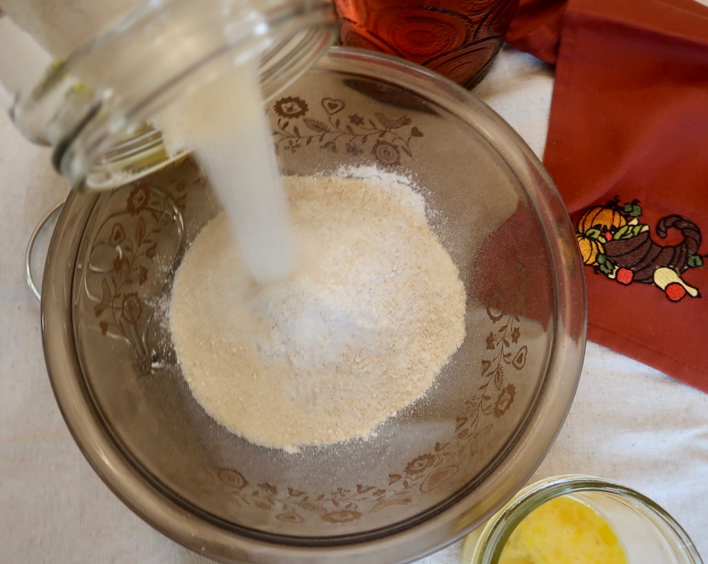 Sugar being poured into a glass bowl containing flour, with a red patterned napkin and a jar of beaten eggs in the background.