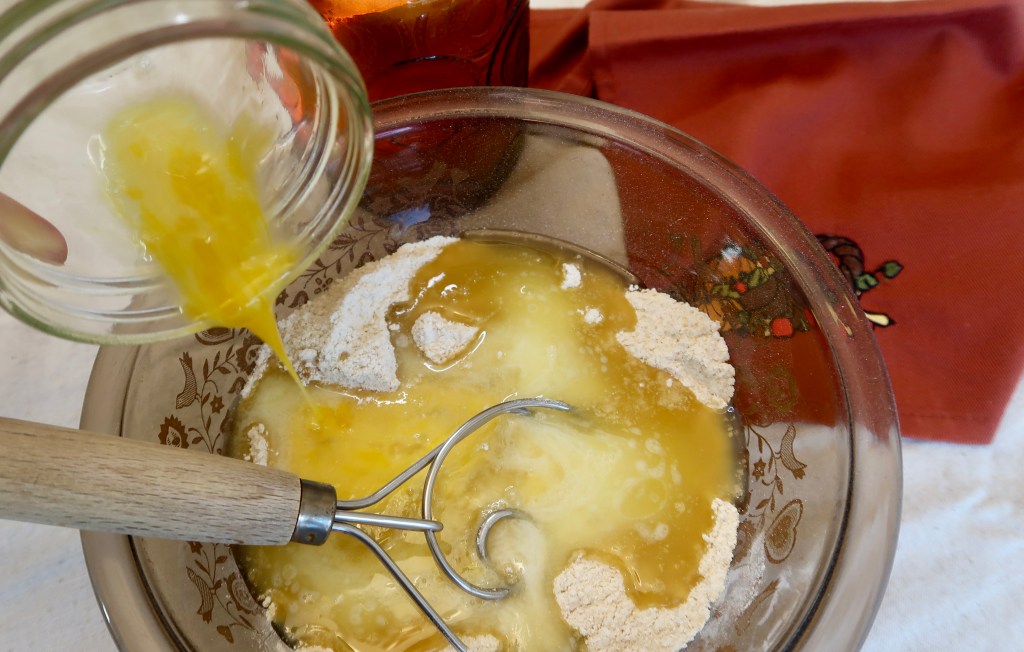 A glass bowl containing a mixture of flour, melted butter, and an egg being poured in from a jar, with a whisk resting inside the bowl and a red cloth in the background.