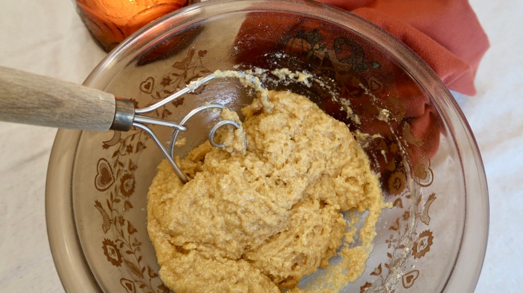 A mixing bowl with creamy cookie dough and a metal mixing attachment, accompanied by a brown cloth in the background.