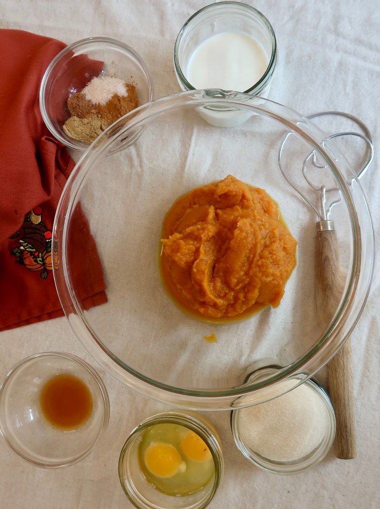 Ingredients for a pumpkin recipe including a bowl of pumpkin puree, milk, eggs, sugar, vanilla extract, and various spices arranged on a beige cloth.