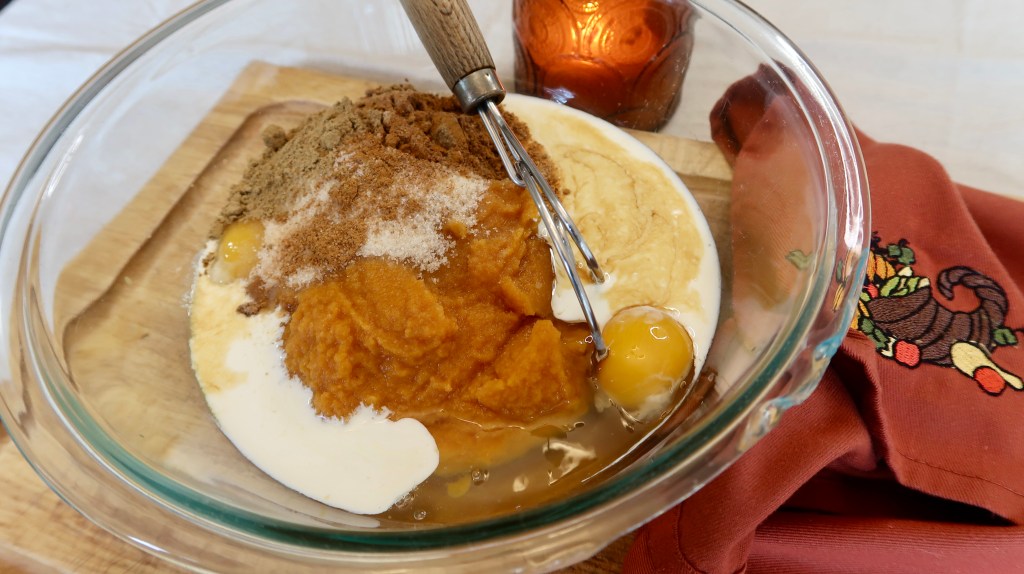 A glass bowl filled with pumpkin puree, eggs, sugar, and spices on a wooden cutting board, with a whisk inside and a decorative cloth nearby.