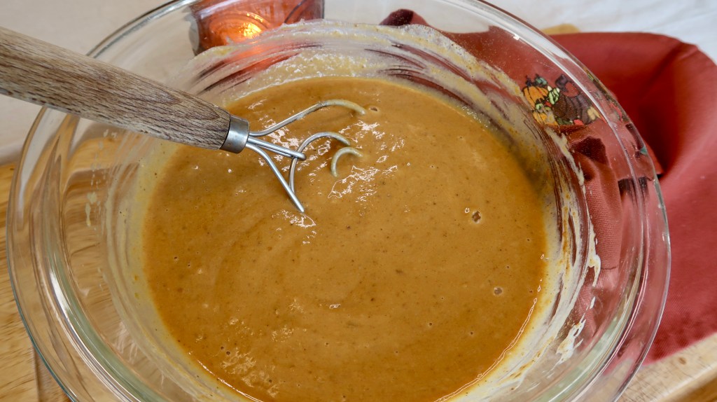 A glass mixing bowl with a smooth, creamy brown batter and a metal whisk, resting on a wooden surface.
