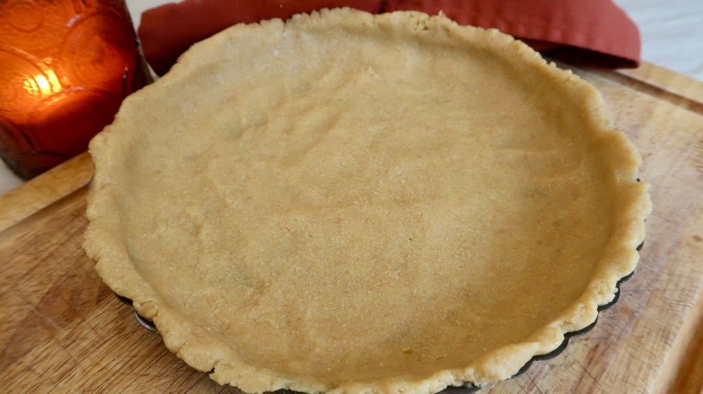 Unbaked pie crust in a pie dish, resting on a wooden surface with a candle and towel in the background.
