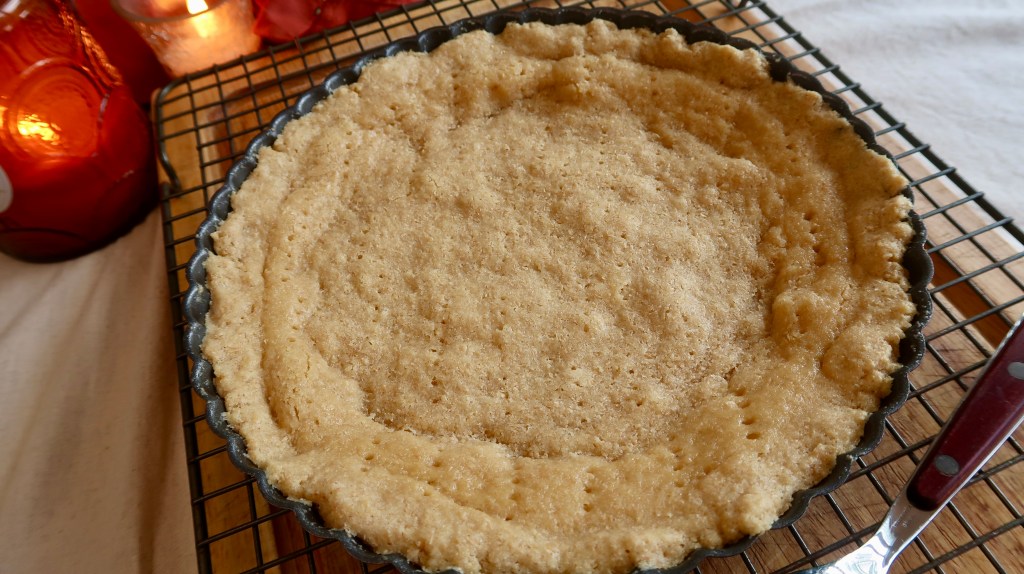 A baked tart crust cooling on a wire rack, with a smooth, light brown surface and fluted edges.