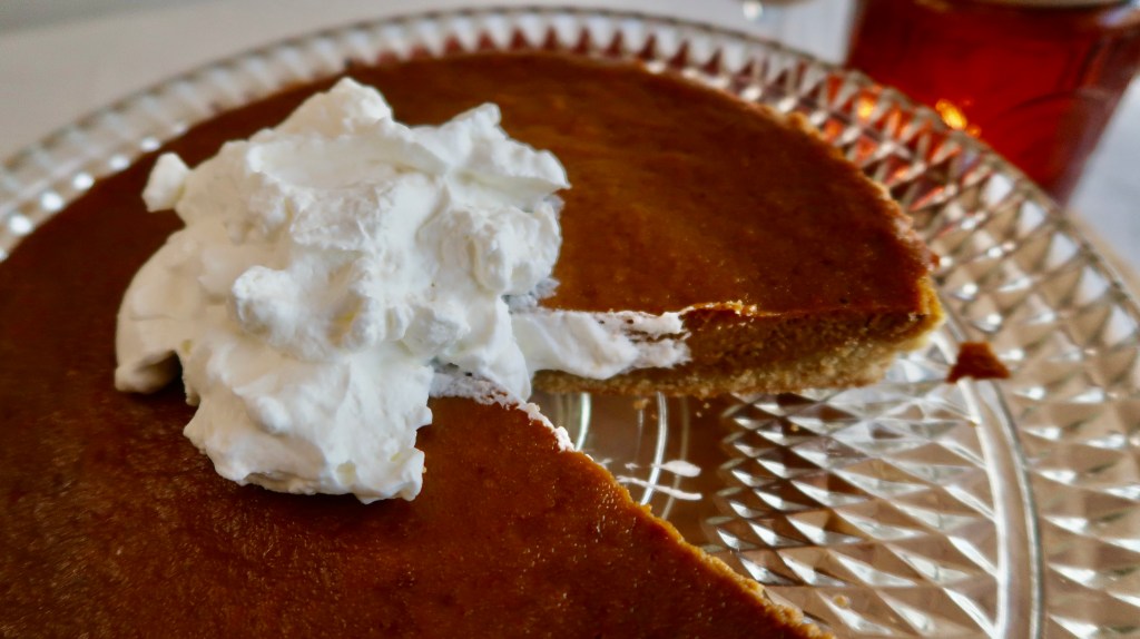 A close-up of a slice of pumpkin pie topped with whipped cream on a decorative glass plate, with a glass of iced tea in the background.