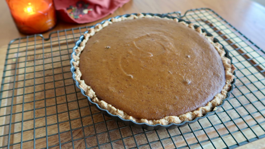 A freshly baked pumpkin pie cooling on a wire rack, with a candle and a decorative cloth in the background.