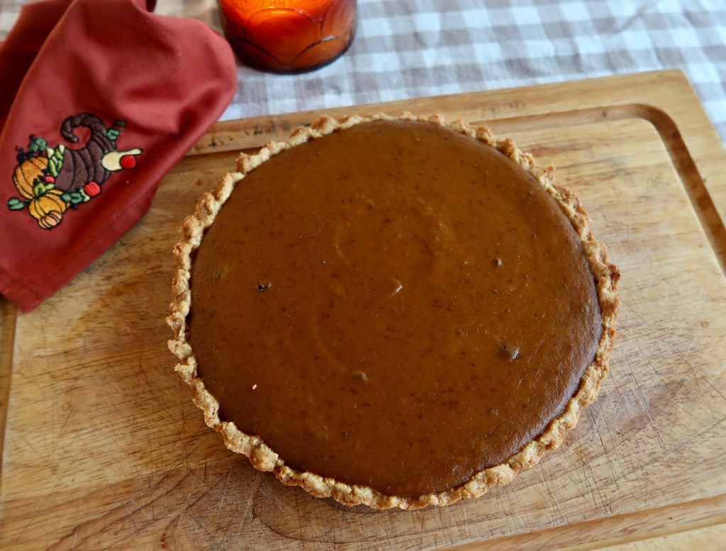 A freshly baked pumpkin pie on a wooden cutting board, featuring a golden-brown crust and smooth, spiced filling. A red cloth napkin with autumn decorations is placed beside the pie, and a candle is in the background on a checkered tablecloth.