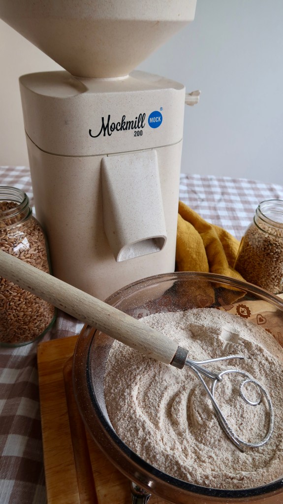 A Mockmill 200 grain mill with grain in jars beside it, and a bowl of freshly ground flour featuring a whisk resting on top.