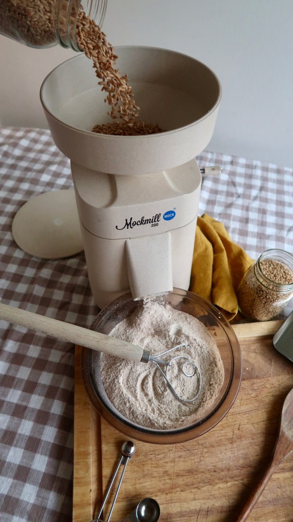 A Mockmill grain mill is shown with whole grains being poured into it, while freshly ground flour sits in a bowl beside it on a wooden cutting board.