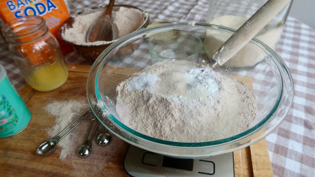 A glass mixing bowl filled with a mixture of flour and baking powder, with a wooden spoon. In the background, there's a jar of baking soda, a bowl of flour, and a container with yellow liquid. The scene is set on a wooden cutting board with measuring spoons and some flour spilled on the surface.