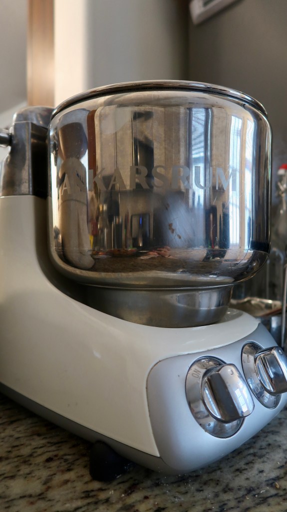 Close-up of a polished stainless steel bowl with 'ANKARSRUM' engraved on it, attached to a white kitchen mixer. The mixer is positioned on a granite countertop.
