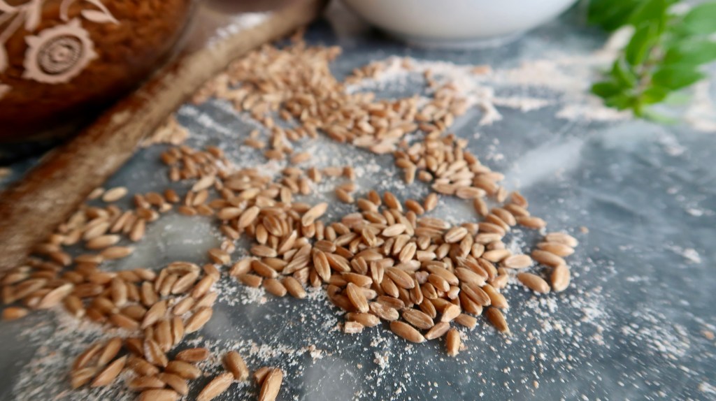 A close-up view of scattered grains on a textured surface, alongside a wooden spoon and a bowl in the background, with some herb leaves visible.