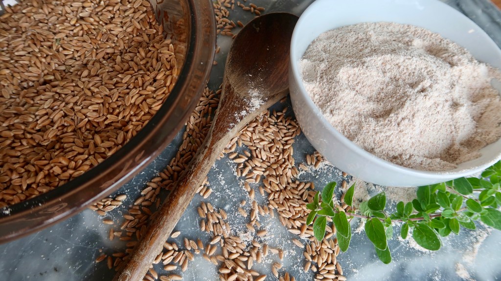 A bowl of whole seeds and a separate bowl of ground flour on a marble surface, accompanied by a wooden spoon and fresh green leaves.