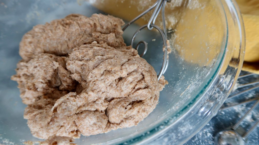 Close-up of thick, brown dough in a glass mixing bowl with a whisk attachment.