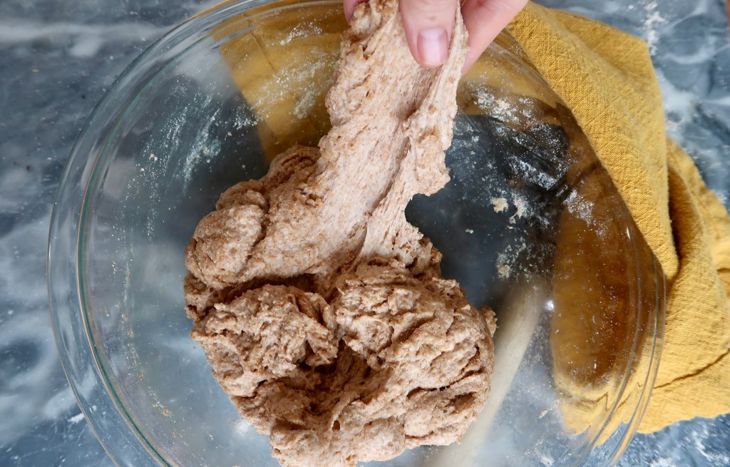 A hand holding a piece of dough above a glass bowl filled with more dough, on a marble countertop with a yellow cloth beside it.