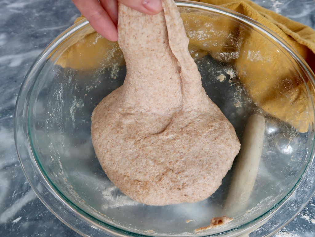 A hand stretches whole wheat dough in a glass bowl, with a rolling pin and a yellow cloth in the background.