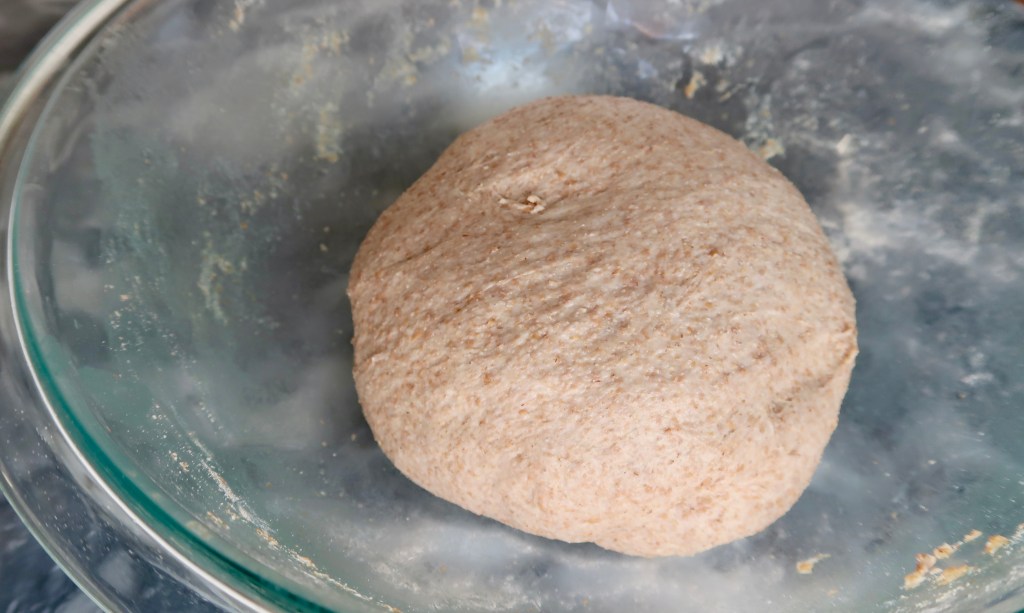 A close-up view of a round ball of whole wheat dough resting in a glass mixing bowl.