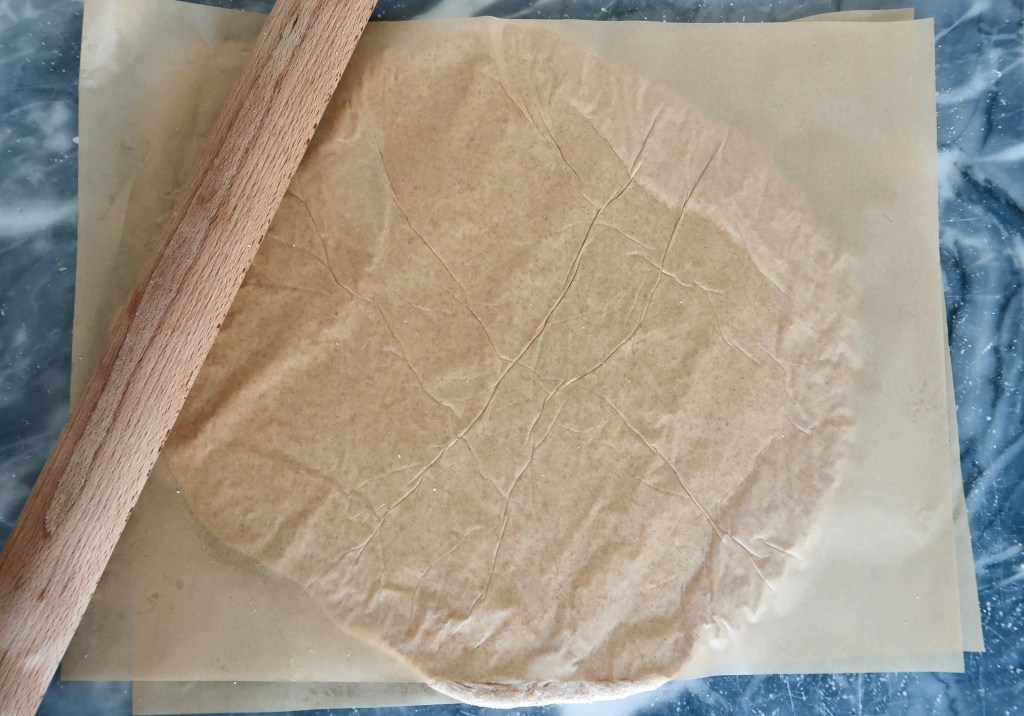 A wooden rolling pin rests on top of a round sheet of parchment paper, with another sheet of parchment below it.