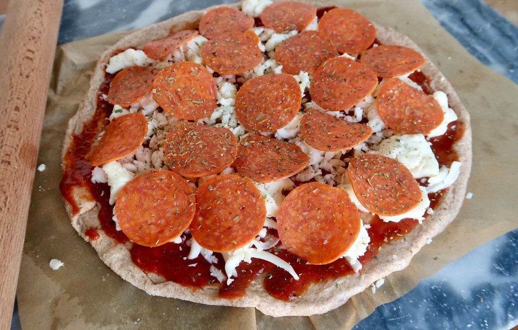 Raw pizza topped with pepperoni, mozzarella cheese, and herbs on a baking paper.