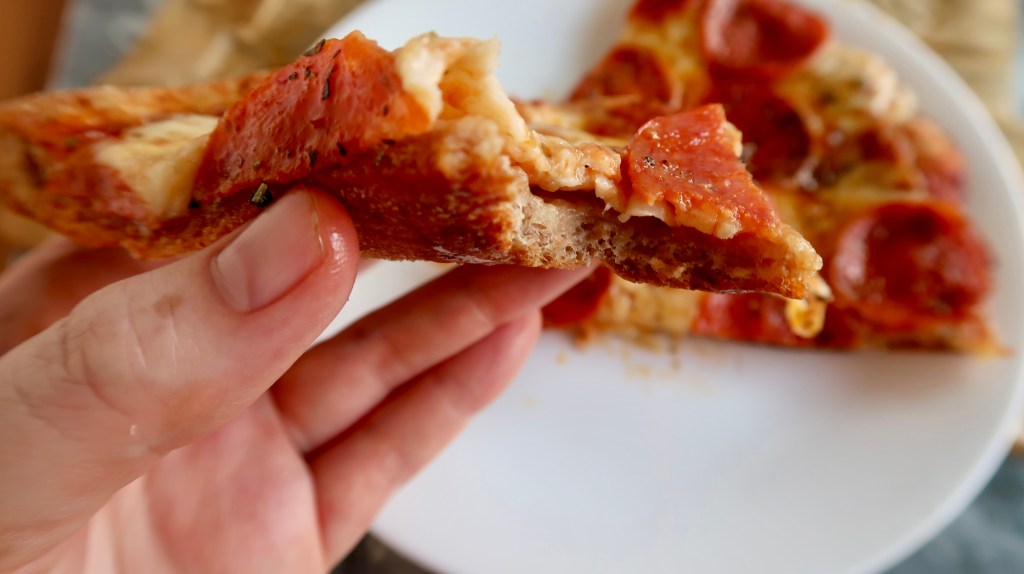 A hand holding a slice of pepperoni pizza, with cheese and toppings clearly visible. A plate of pizza slices is in the background.