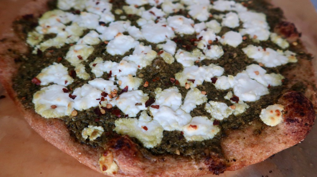 Close-up of a freshly baked pizza topped with pesto, dollops of ricotta cheese, and red pepper flakes on a crispy crust.