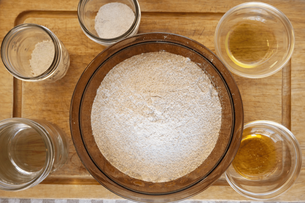 A wooden board displaying a brown bowl filled with flour, surrounded by small glass jars containing different dry ingredients and liquids.