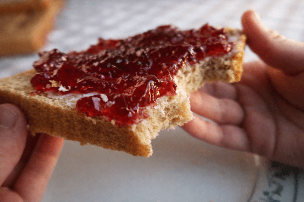 A close-up of a hand holding a slice of bread spread with red jelly, with a bite taken out of it.