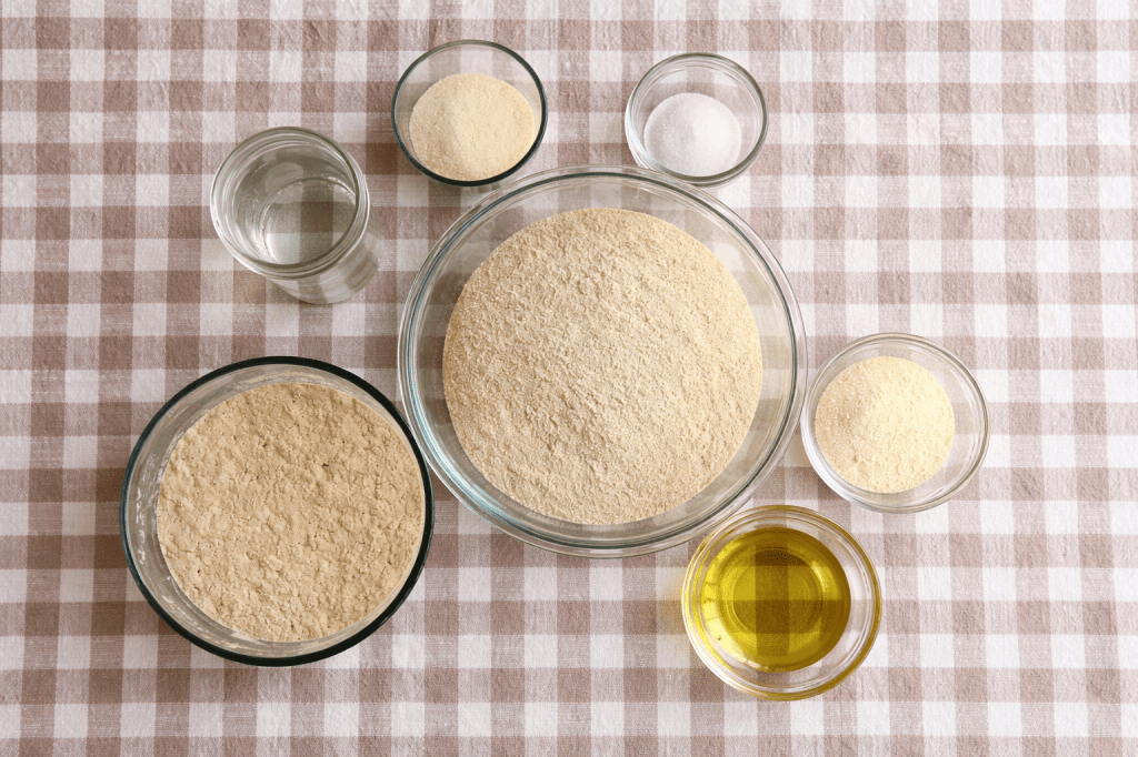 A flat lay arrangement of various ingredients on a checkered tablecloth, including bowls of flour, oil, sugar, and a glass of water.