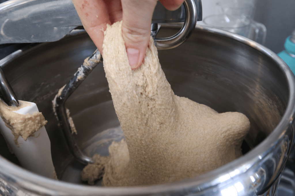 A hand holding a thick, sticky mixture of dough or batter being pulled from a mixing bowl with a mixing attachment.