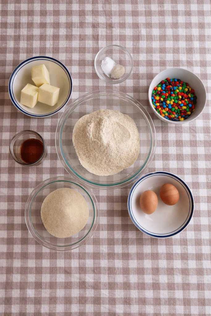 A variety of baking ingredients arranged on a checkered tablecloth, including butter, sugar, flour, eggs, vanilla extract, baking powder, and colorful candy pieces.