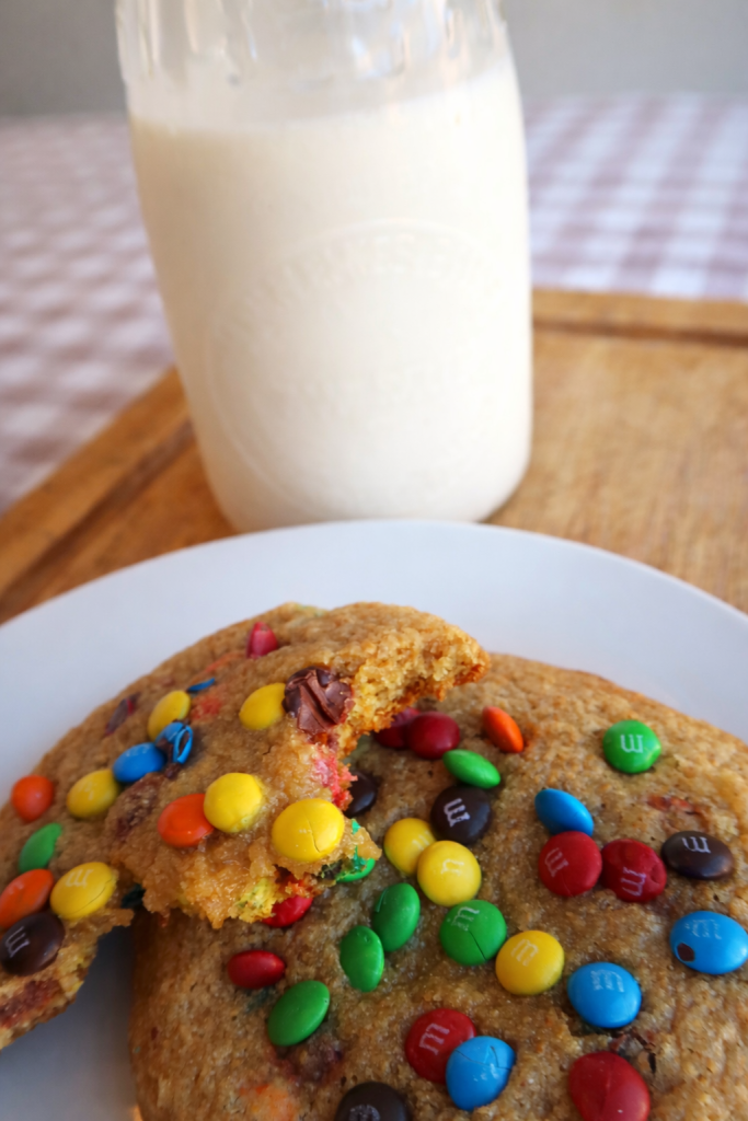 Close-up of two large cookies with colorful chocolate candies, one with a bite taken out, alongside a mason jar of milk on a wooden surface.