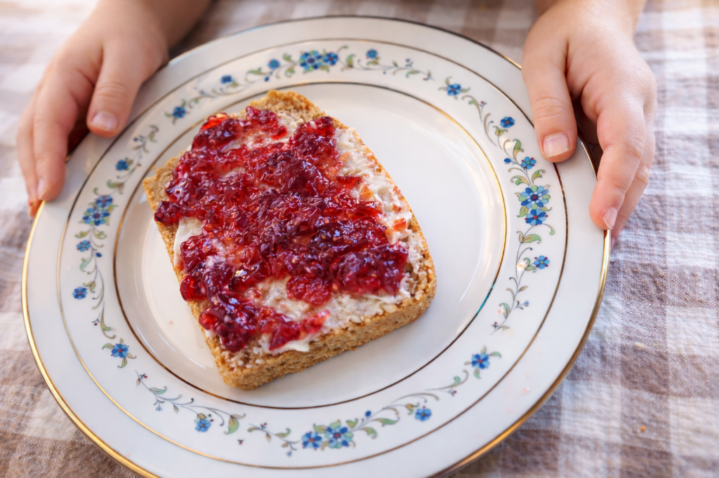 A child's hands holding a plate with a slice of bread topped with cream cheese and strawberry jam, placed on a decorative floral plate, on a checkered tablecloth.