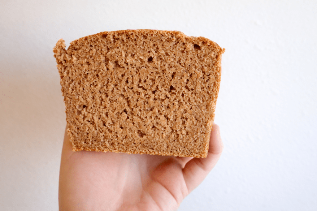 A person holding a slice of freshly baked brown bread, showcasing the texture and crumb structure against a plain white background.