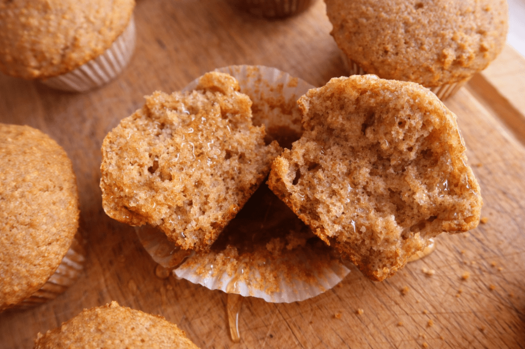A close-up of a freshly baked muffin, partially peeled from its wrapper, showing a moist and fluffy interior, with additional muffins in the background.