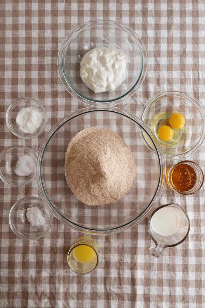 Top view of various baking ingredients arranged on a checkered tablecloth, including a bowl of brown sugar, eggs, yogurt, and liquid ingredients.