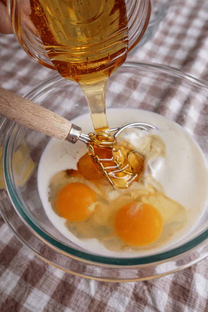 A glass bowl containing three raw egg yolks, milk, and a whisk, with honey being poured into the mixture from a jar.